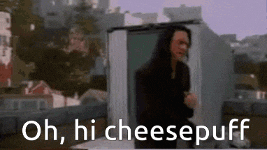 a man with long hair is standing in front of a building with the words `` oh , hi cheesepuff '' written below him .