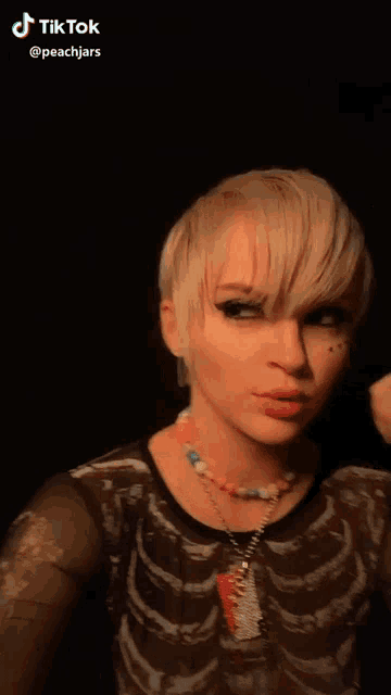 a woman with short blonde hair wearing a necklace and a skeleton shirt .