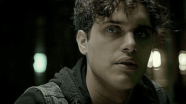 a close up of a man 's face with curly hair wearing a black shirt and a denim vest .