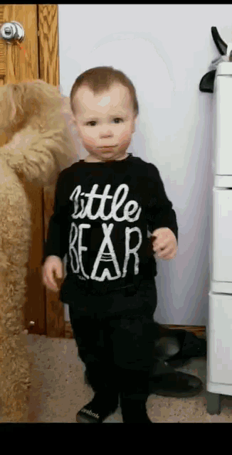 a toddler wearing a little bear shirt stands in front of a teddy bear