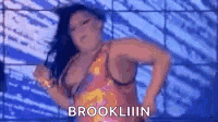 a woman in a pink dress is dancing in front of a blue wall and says brookliiin .