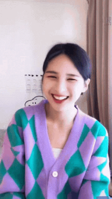 a woman wearing a purple and green cardigan is smiling and looking at the camera .