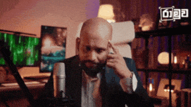a bald man with a beard is sitting in front of a microphone and touching his ear .