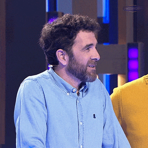 a man with a beard is wearing a blue shirt with a black embroidered logo on the left sleeve