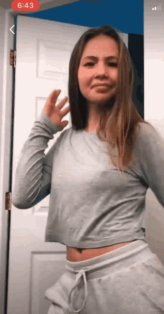 a woman in a grey crop top and white sweatpants is standing in front of a door with the time 6:43