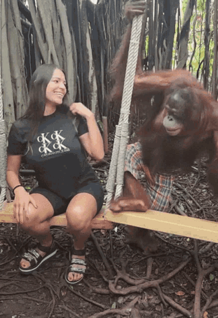 a woman wearing a black ck & k shirt sits next to an orangutan