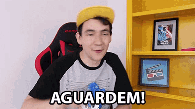 a young man wearing a yellow hat is sitting in a red and black gaming chair and says aguardem