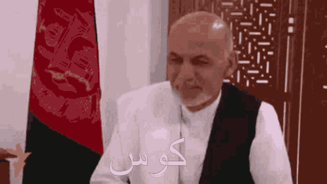 a bald man with a beard is sitting in front of a flag with arabic writing .