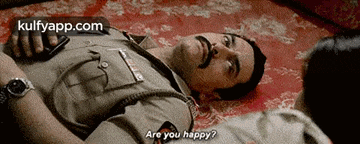 a man in a police uniform is laying on the floor while a woman looks on and says " are you happy "