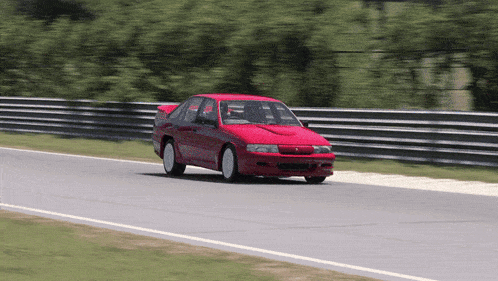 a red car is driving down a road with a fence in the background