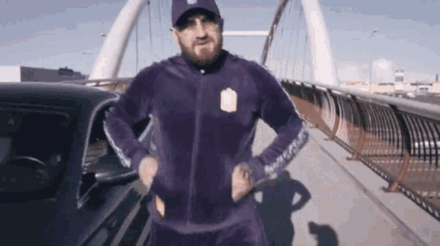a man in a purple tracksuit is standing in front of a car on a bridge .