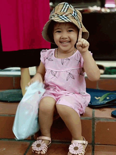 a little girl wearing a plaid hat and a pink dress is smiling and pointing upwards