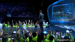 a man in a suit stands on a stage in front of a crowd of people with their hands in the air