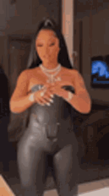 a woman in a black leather jumpsuit is standing in a room with her hands on her chest .