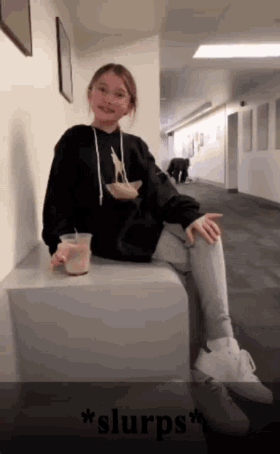 a girl in a black hoodie sits on a cube in a hallway holding a cup of coffee