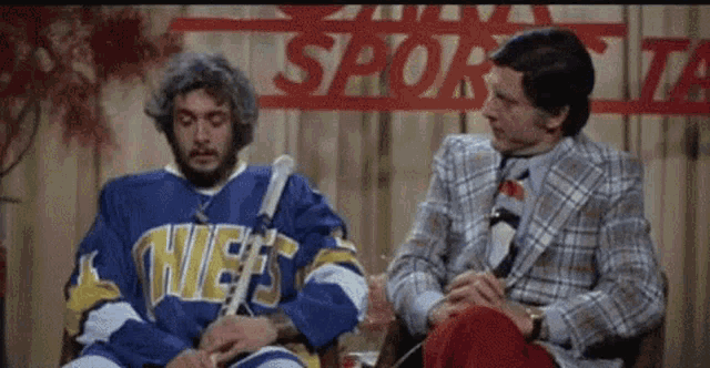 a man in a chiefs jersey sits next to a man in a suit
