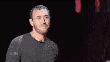a man with a beard is standing in front of a microphone in a dark room and making a funny face .