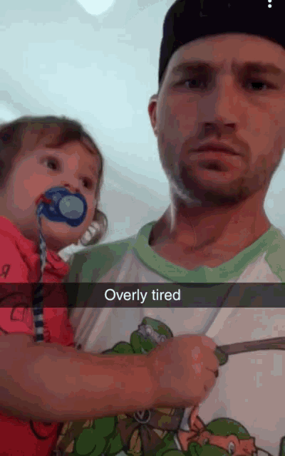 a man is holding a baby with a pacifier in his mouth and overly tired is written on the bottom