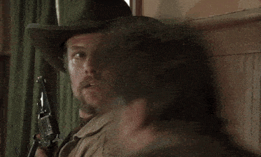 a man with a beard wearing a cowboy hat holds a gun