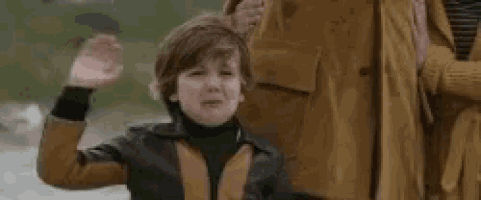 a young boy is waving his hand in the air while standing next to a woman in a brown coat .