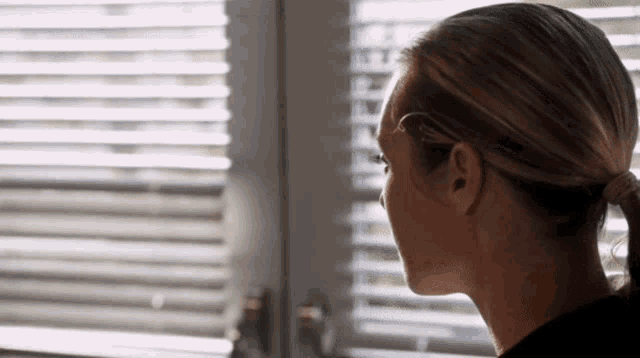 a woman in a ponytail is looking out of a window with blinds .