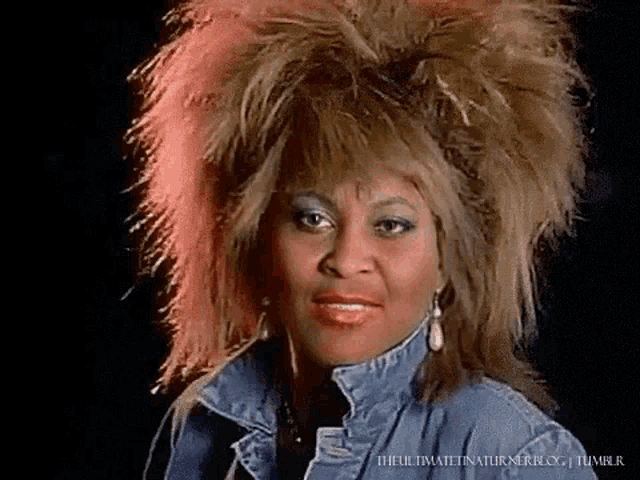 a woman wearing a denim jacket and earrings has a very large hairdo .