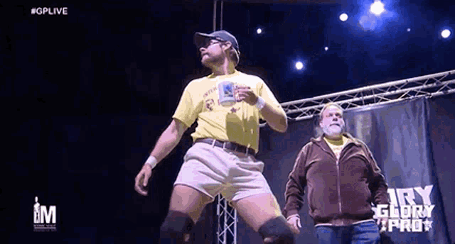 a man in a yellow shirt is holding a cup while a man in a brown jacket watches .