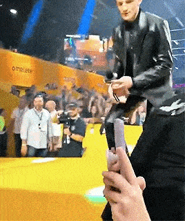 a man in a leather jacket is standing on a stage holding a microphone while a person holds a cell phone .