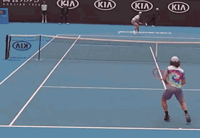 a man in a tie dye shirt stands on a kia tennis court