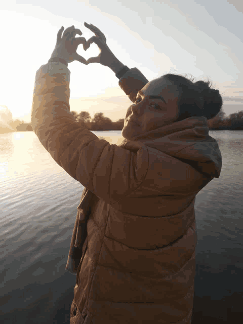a woman making a heart shape with her hands near a body of water