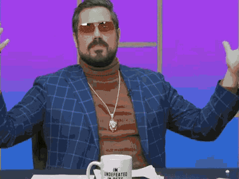 a man wearing sunglasses and a turtleneck is sitting at a table with a mug that says im undefeated