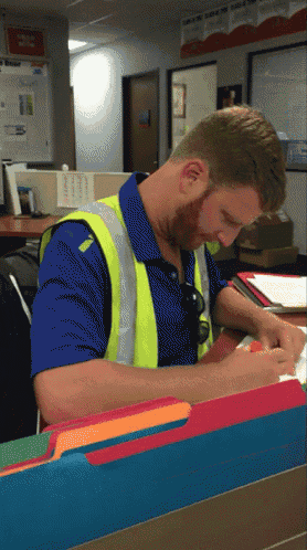a man wearing a blue shirt and a yellow vest sits at a desk looking at a piece of paper
