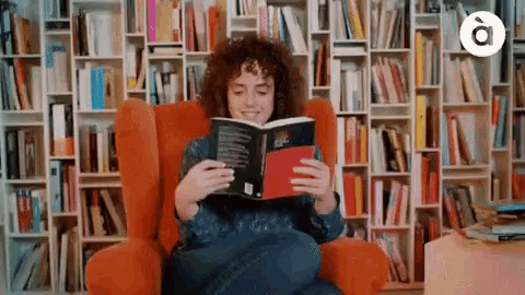 a woman sits in an orange chair reading a book in front of bookshelves