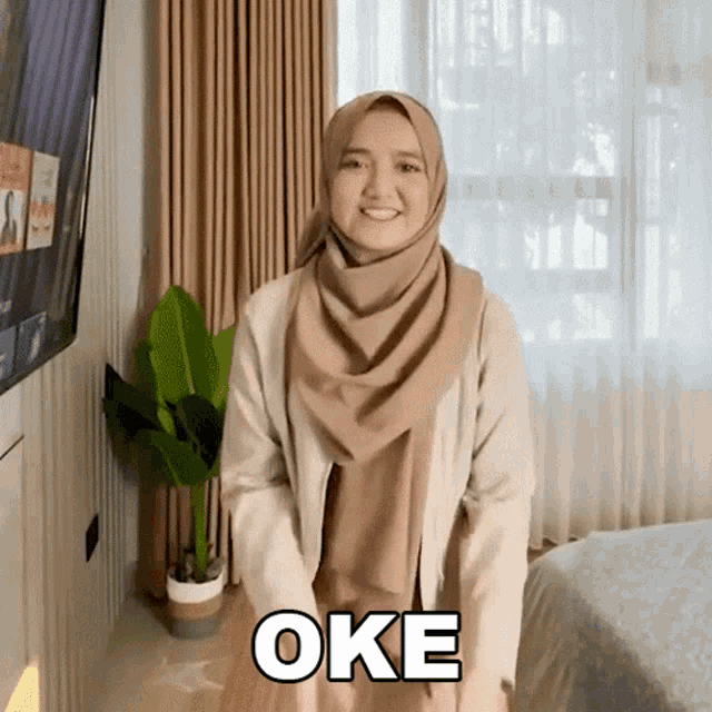a woman wearing a hijab is standing in front of a television and says oke