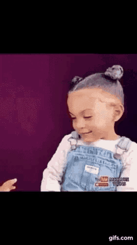 a little girl in overalls is making a funny face while standing in front of a purple wall .