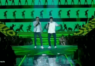 two men singing into microphones on a stage in front of a green background