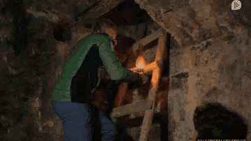 a man in a green jacket is standing in a cave with a ladder and a light .