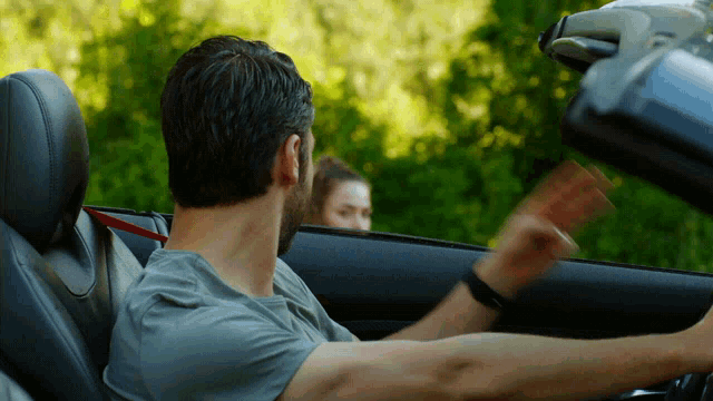 a man in a grey shirt is driving a car with a woman behind him