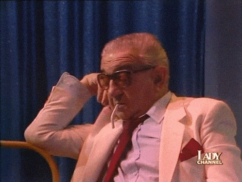 a man in a white suit and red tie is sitting in front of a lady channel sign