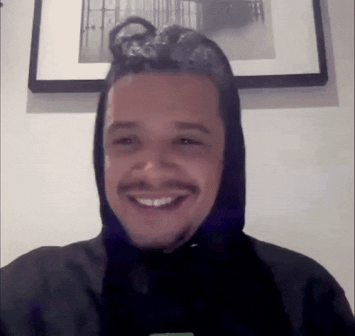 a man wearing a black hoodie is smiling in front of a framed picture on a wall .