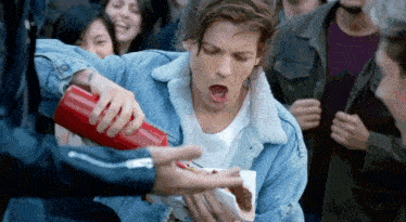 a man in a denim jacket is eating a hot dog while a man pours ketchup on him .