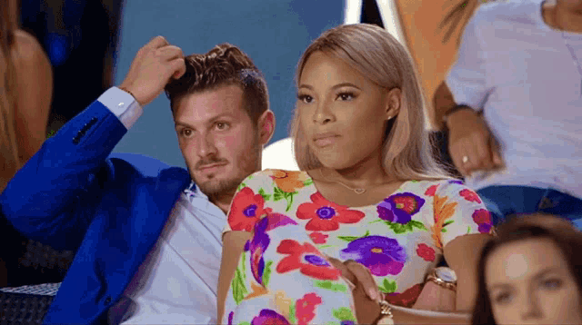 a man in a blue suit scratches his head while a woman in a floral dress sits next to him