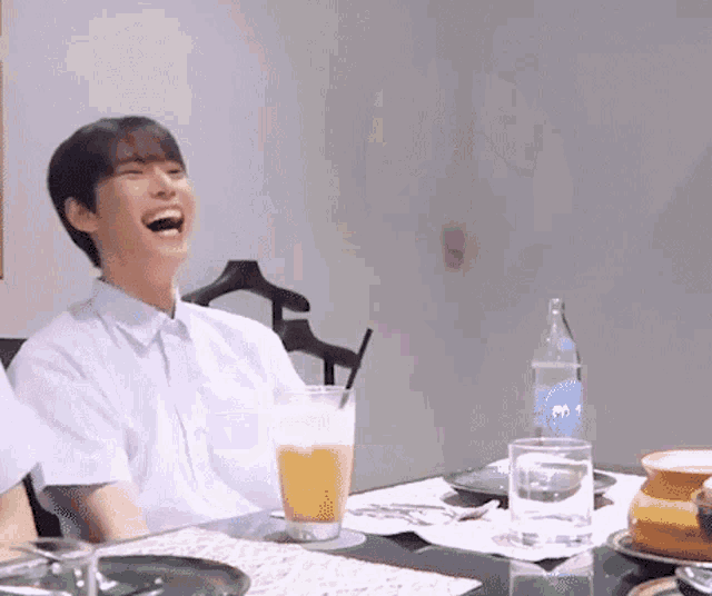 a man is laughing while sitting at a table with a glass of orange juice and a bottle of water .