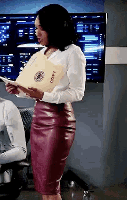 a woman in a white shirt and a red leather skirt is standing in front of a television holding a folder .