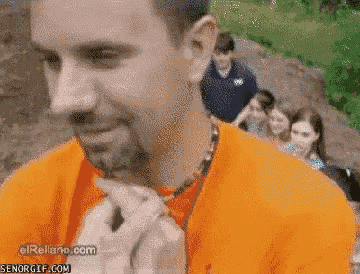 a man in an orange shirt is eating a sandwich while a group of people are standing behind him
