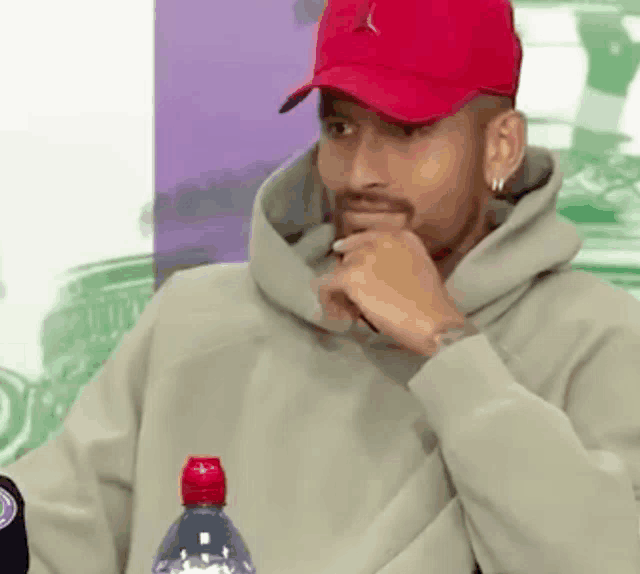 a man wearing a red hat and a hoodie is sitting at a table with his hand on his chin .