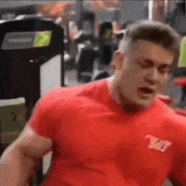 a man in a red shirt is flexing his muscles in a gym