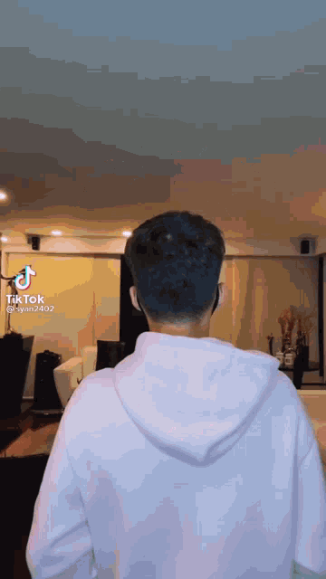 a man wearing headphones and a white hoodie is standing in a living room with tik tok written on the bottom