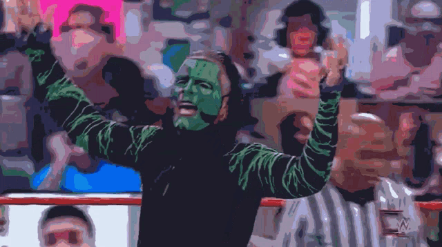 a man with green paint on his face stands in front of a crowd with his arms in the air
