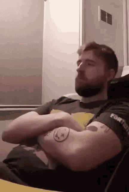 a man with a beard and a tattoo on his arm is sitting with his arms crossed on a couch .